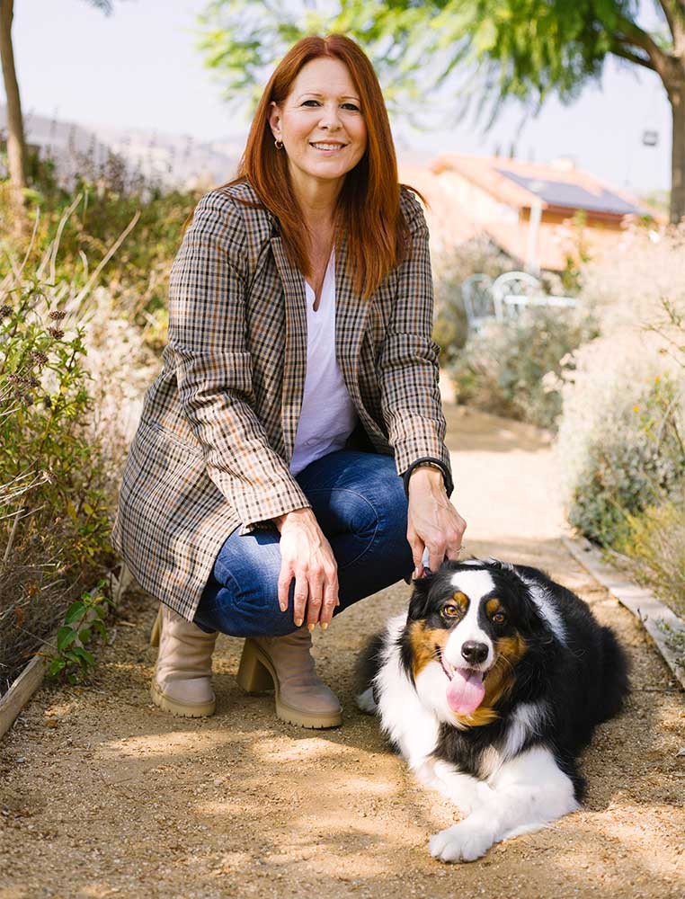 A woman kneeling next to a dog on a trail, smiling at the camera